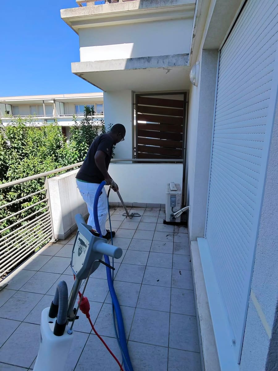 Soum passe le suceur d'injection-extraction sur le carrelage d'un balcon, volet roulant blanc et garde-corps