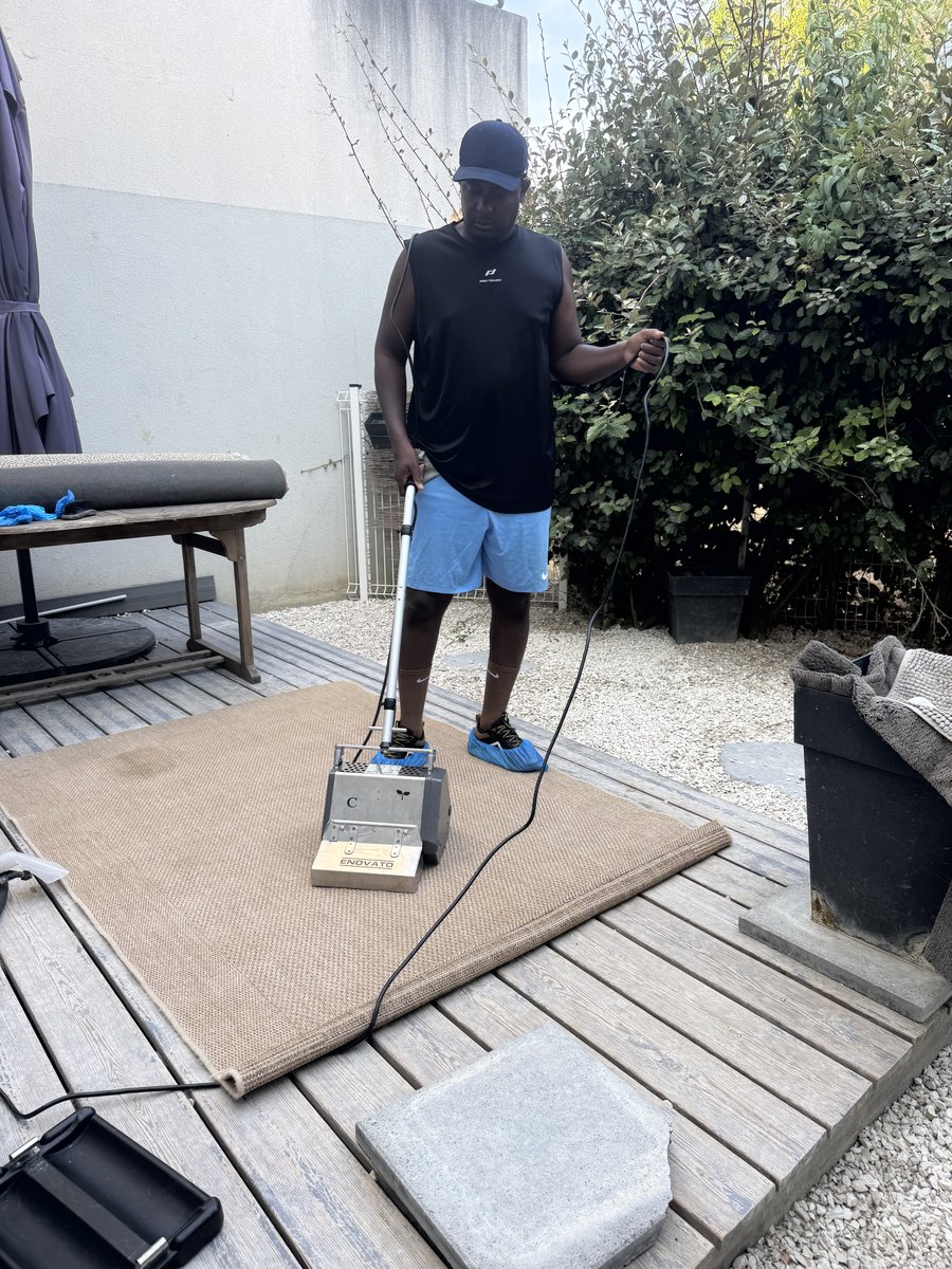 Soum passe la brosse rotative sur un grand tapis clair déroulé sur une terrasse en bois