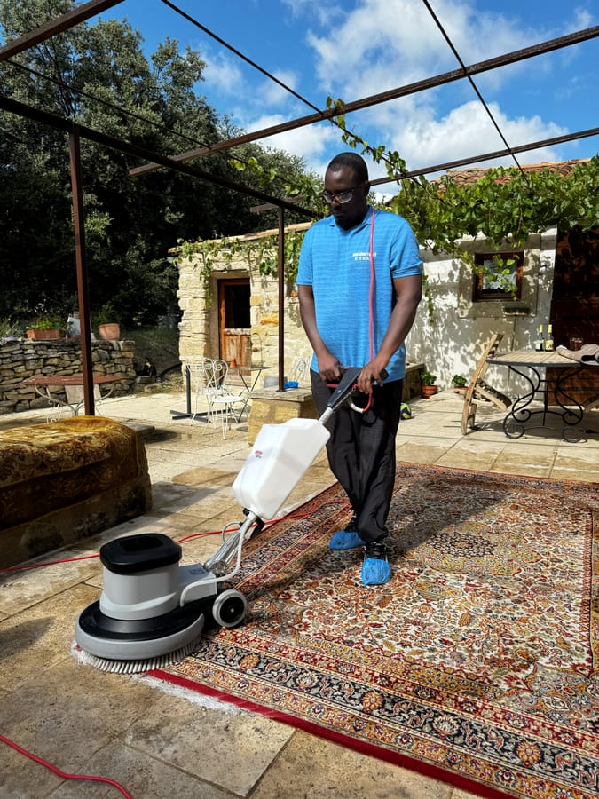 Soum passe la monobrosse sur un tapis à motifs sous une pergola, mas en pierre au fond