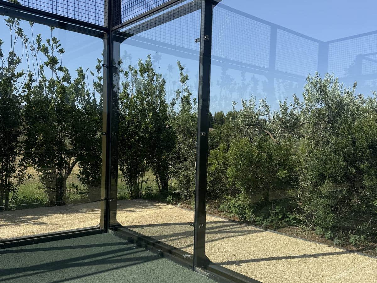 Parois vitrées d’un court de padel, structure noire et gazon synthétique, arbres au fond