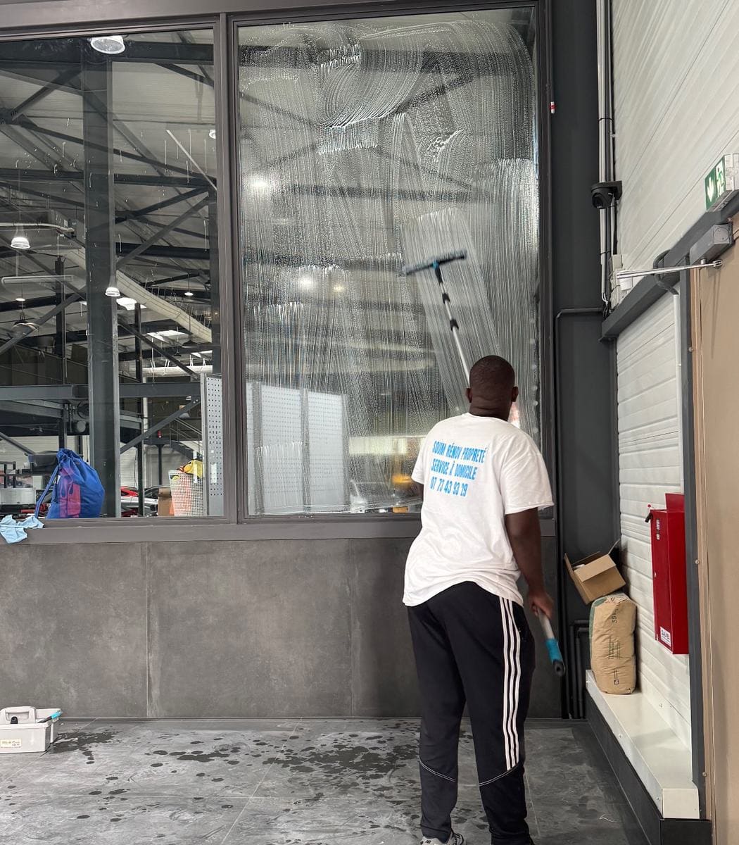 Soum passe la raclette sur une grande vitre ruisselante d’eau savonneuse dans un local professionnel