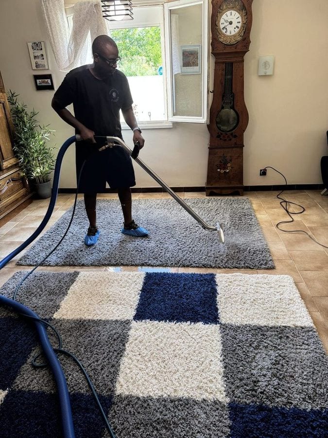 Soum nettoie à la buse un tapis à damier bleu et blanc posé sur un parquet