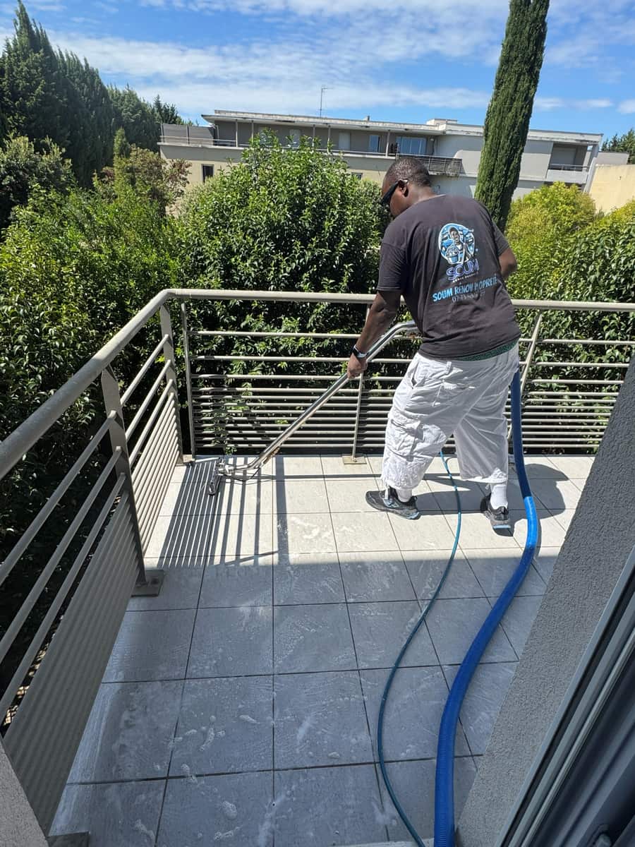 Soum nettoie le carrelage d'un balcon au suceur d'injection-extraction, garde-corps métallique et verdure autour