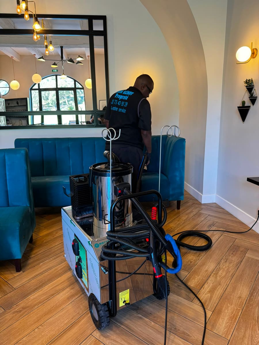 Soum, de dos, passe le nettoyeur vapeur derrière les banquettes turquoise d'une salle, machine inox au premier plan