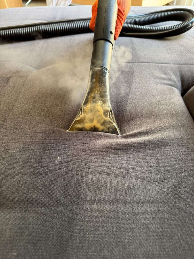 Gros plan du suceur vapeur sur l'assise d'un canapé en tissu gris sombre, la vapeur est visible