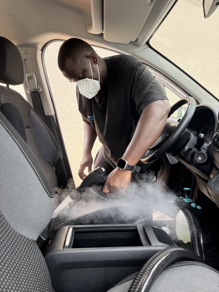 Soum, masqué, passe le nettoyeur vapeur sur la console centrale d'une voiture, la vapeur est visible