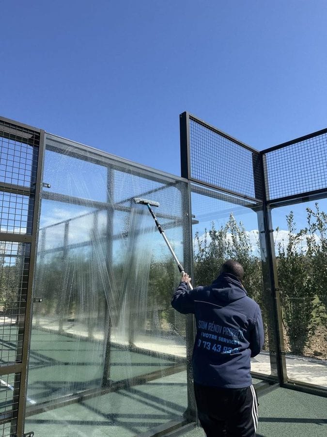 Soum lave à la perche le vitrage d'un terrain de padel, vu de l'extérieur