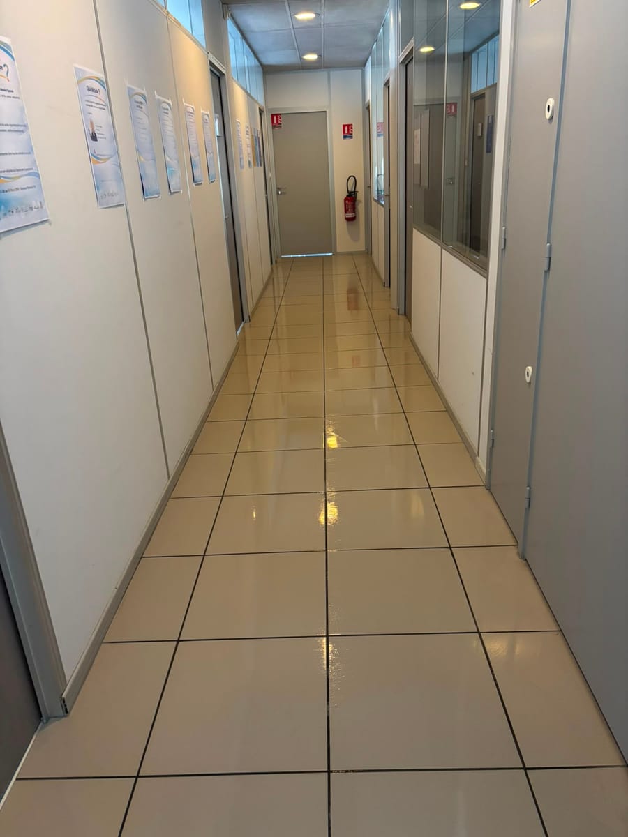 Couloir de bureaux au carrelage clair lavé, cloisons vitrées et affiches le long du mur