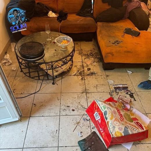 Salon dont le sol est jonché de détritus autour d’un canapé orange et d’une table basse