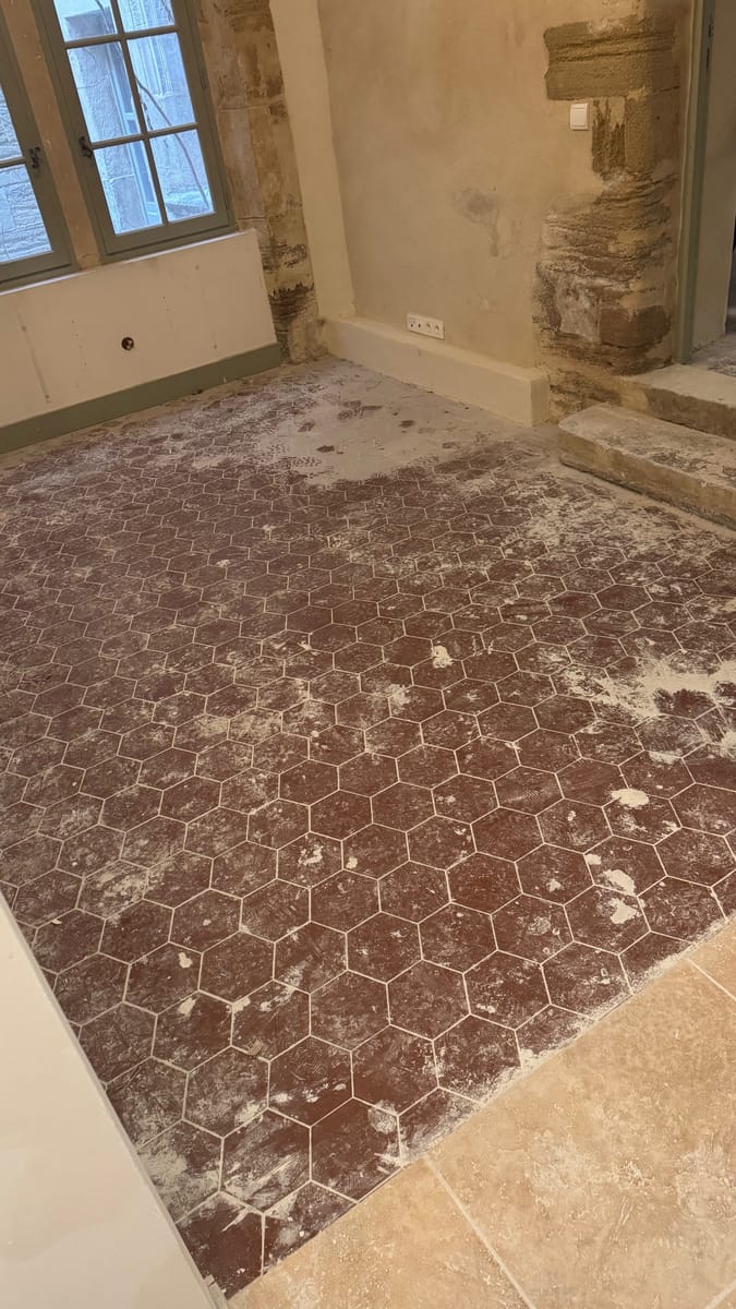 Tomettes hexagonales rouges couvertes de poussière de chantier dans une pièce aux murs de pierre