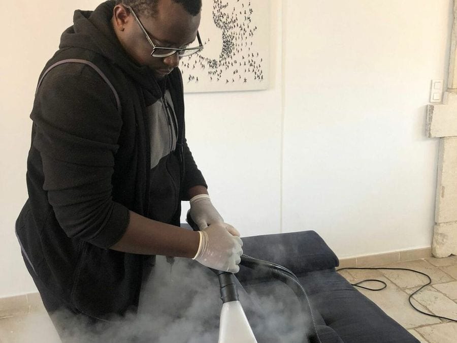 Soum passe le nettoyeur vapeur sur une méridienne en tissu sombre, la vapeur est visible